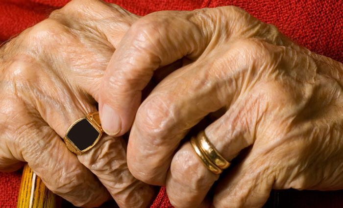 Gerimpelde handen van een oude man. Hij draagt 2 trouwringen en een zegelring. Hij houdt een wandelstok in zijn handern en draagt een rode trui.