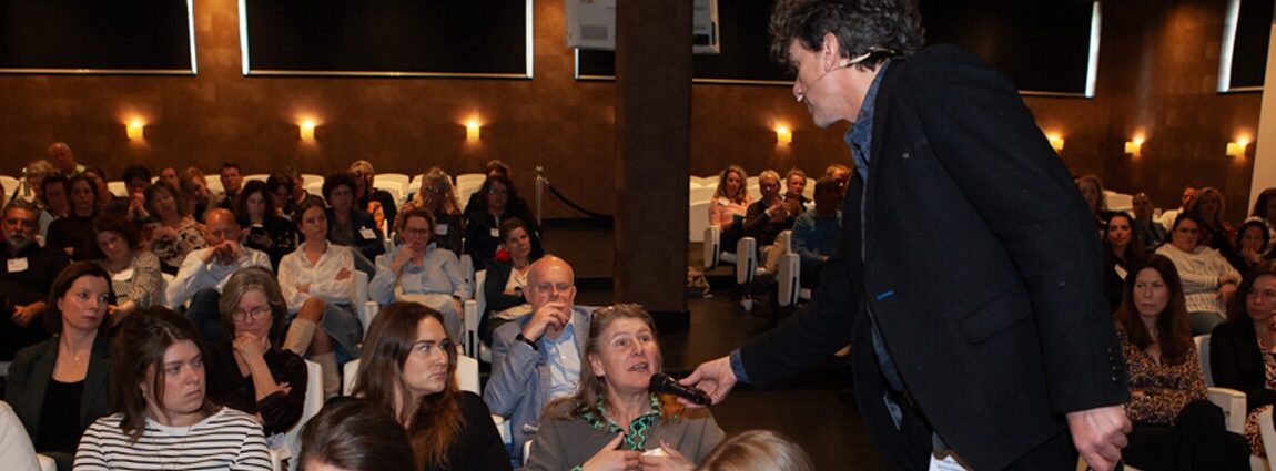 Overzicht van de zaal met leden. Een spreker interviewt iemand in de zaal.