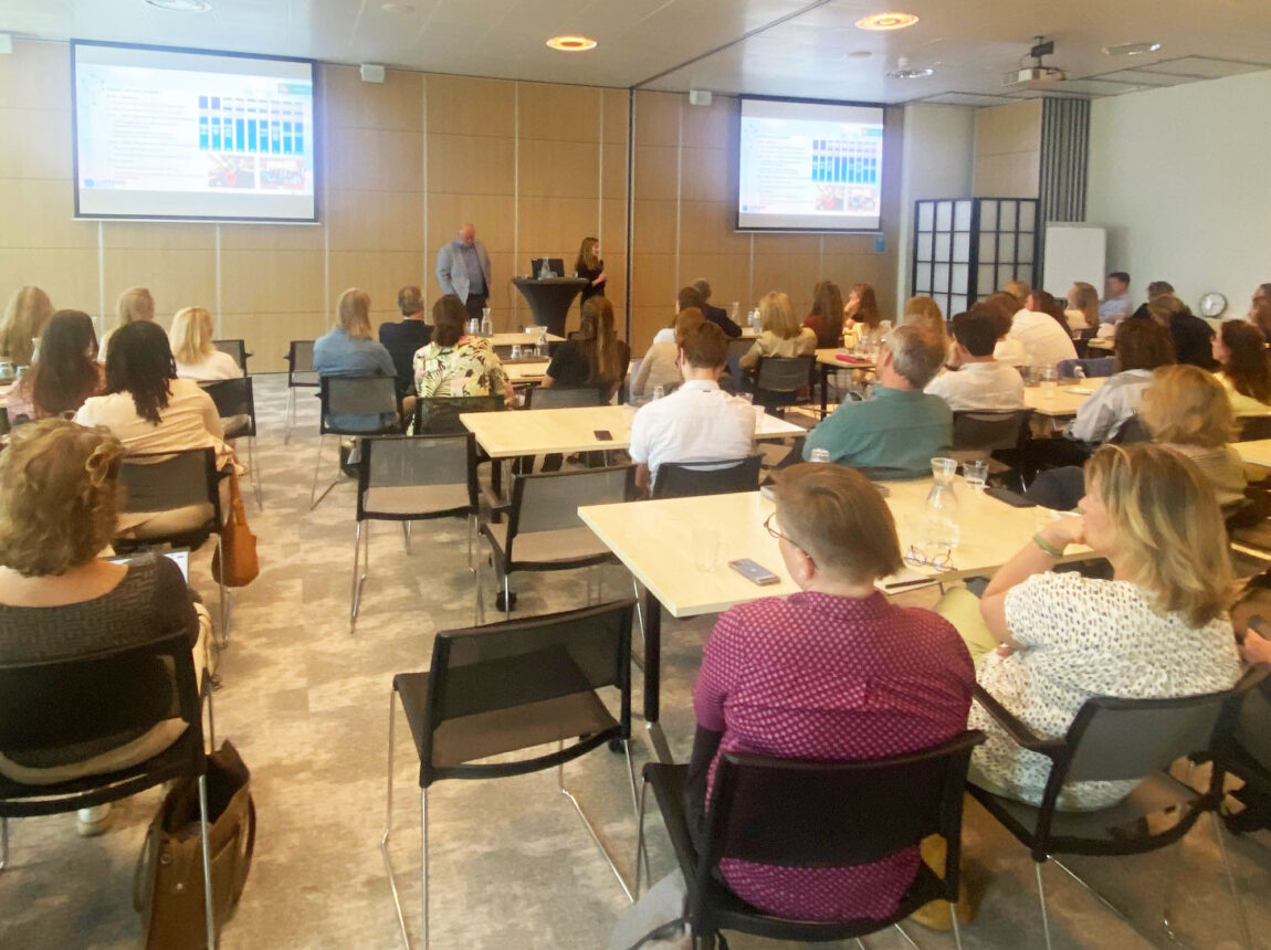 Foto die genomen is tijdens de bijeenkomst. Mensen zitten in een zaal te kijken en luisteren naar een presentatie.