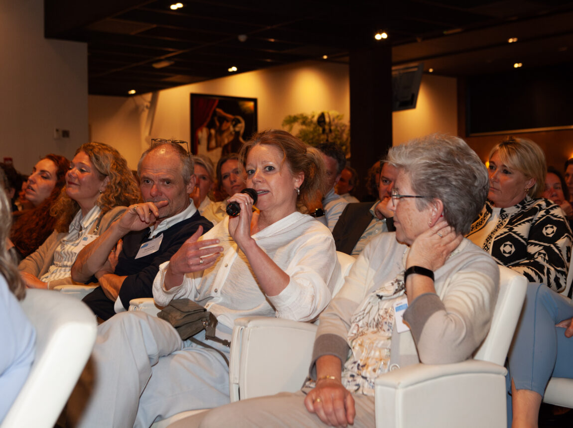 Congreszaal vol mensen waarvan een vrouw door een microfoon praat.