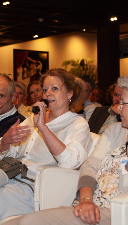 Congreszaal vol mensen waarvan een vrouw door een microfoon praat.