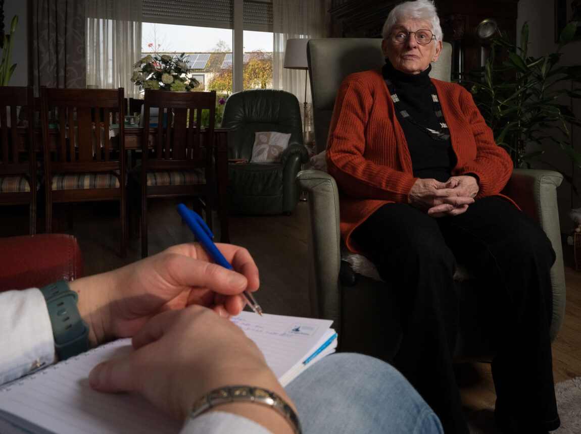 Man maakt aantekeningen op een notitieblok bij een oudere vrouw thuis.
